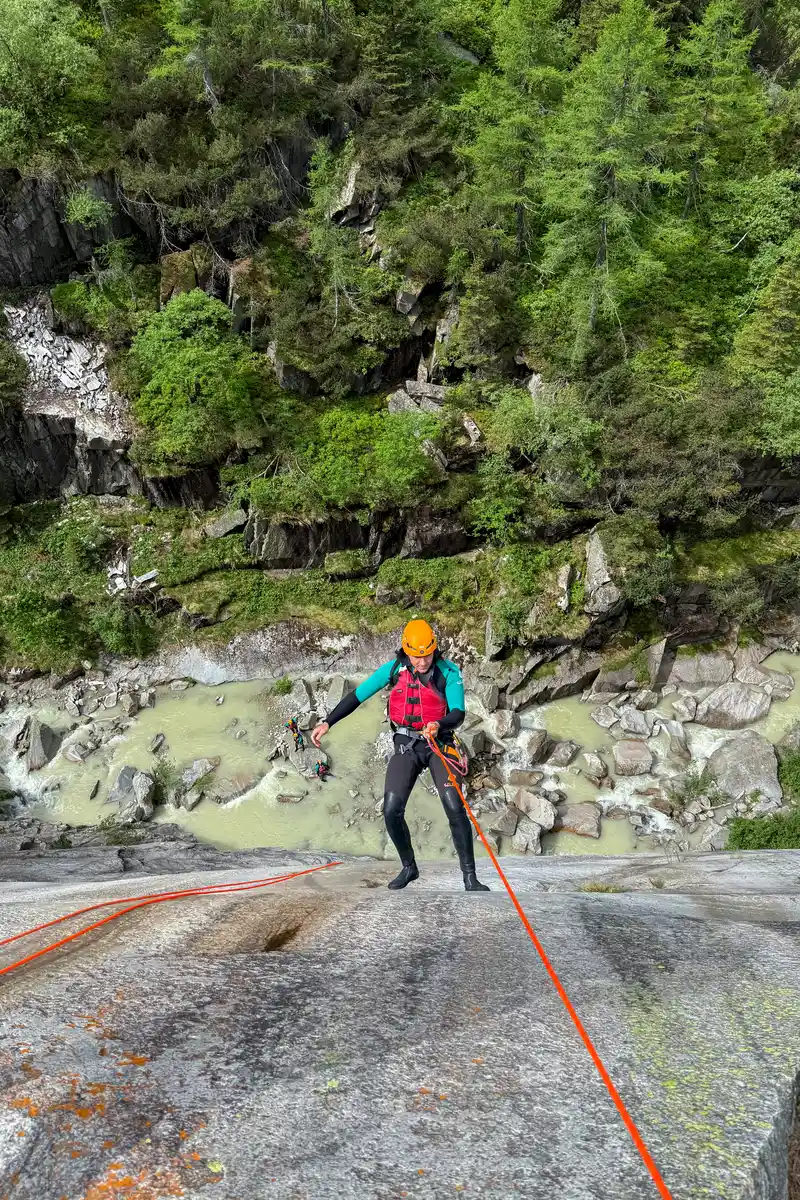 A canyoner in an orange helmet rappelling down a granite wall above a glacial river
